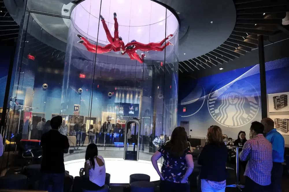 Two people in red suits indoor skydiving in a glass wind tunnel watched by spectators.