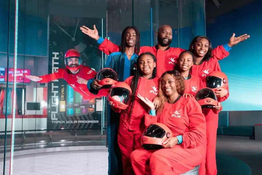 Group of people in red jumpsuits pose before an indoor skydiving tunnel with a flyer.