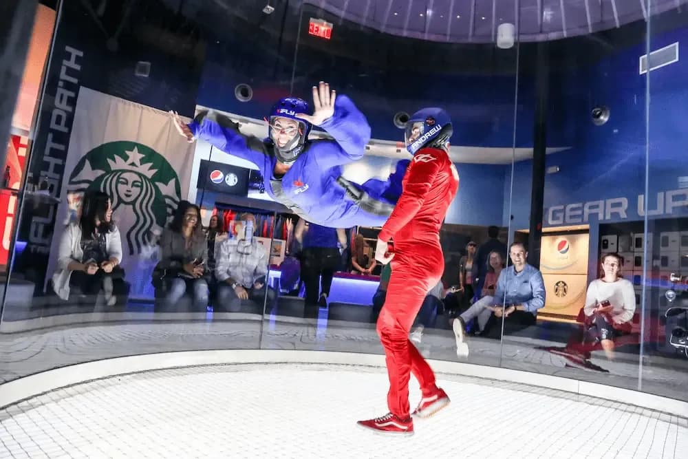 Two people in flight suits, one flying in a wind tunnel, the other guiding.