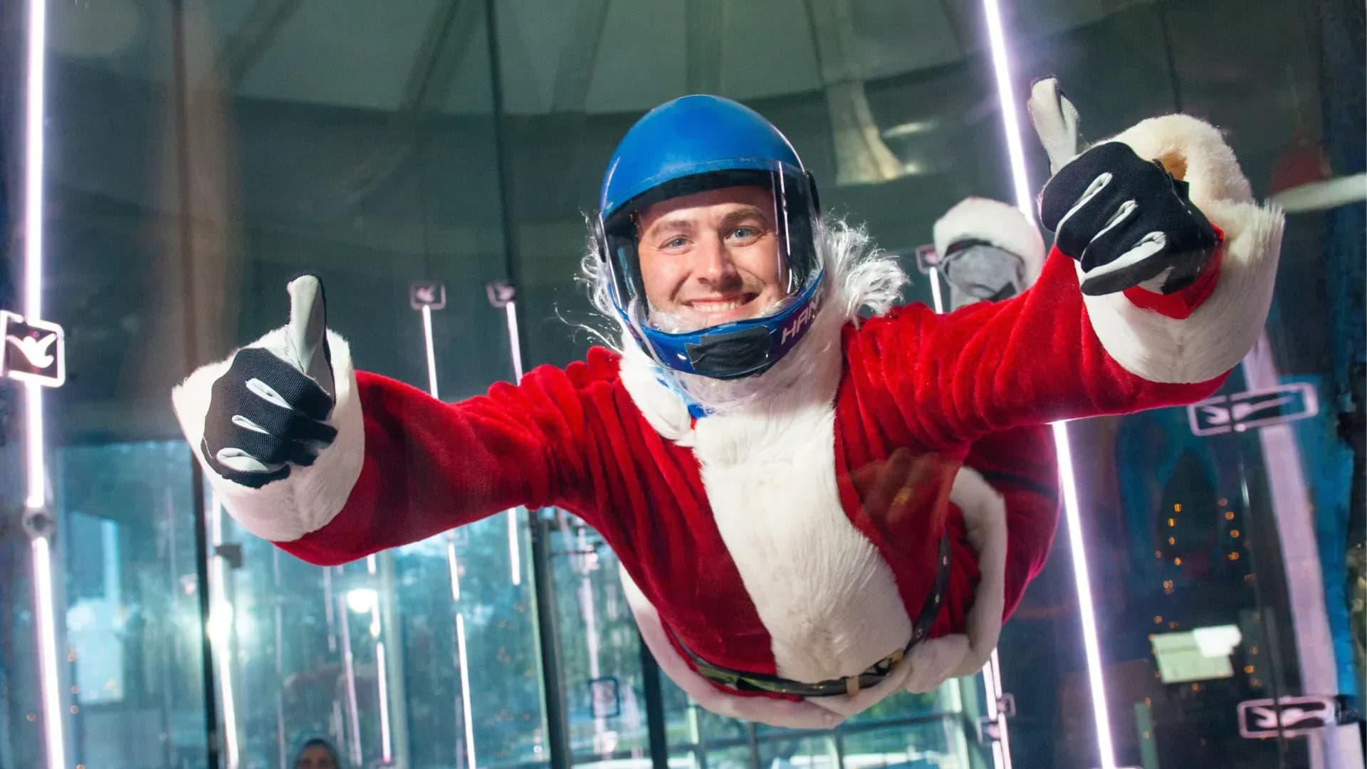 Santa Claus in a blue helmet smiles, giving thumbs up while indoor skydiving.