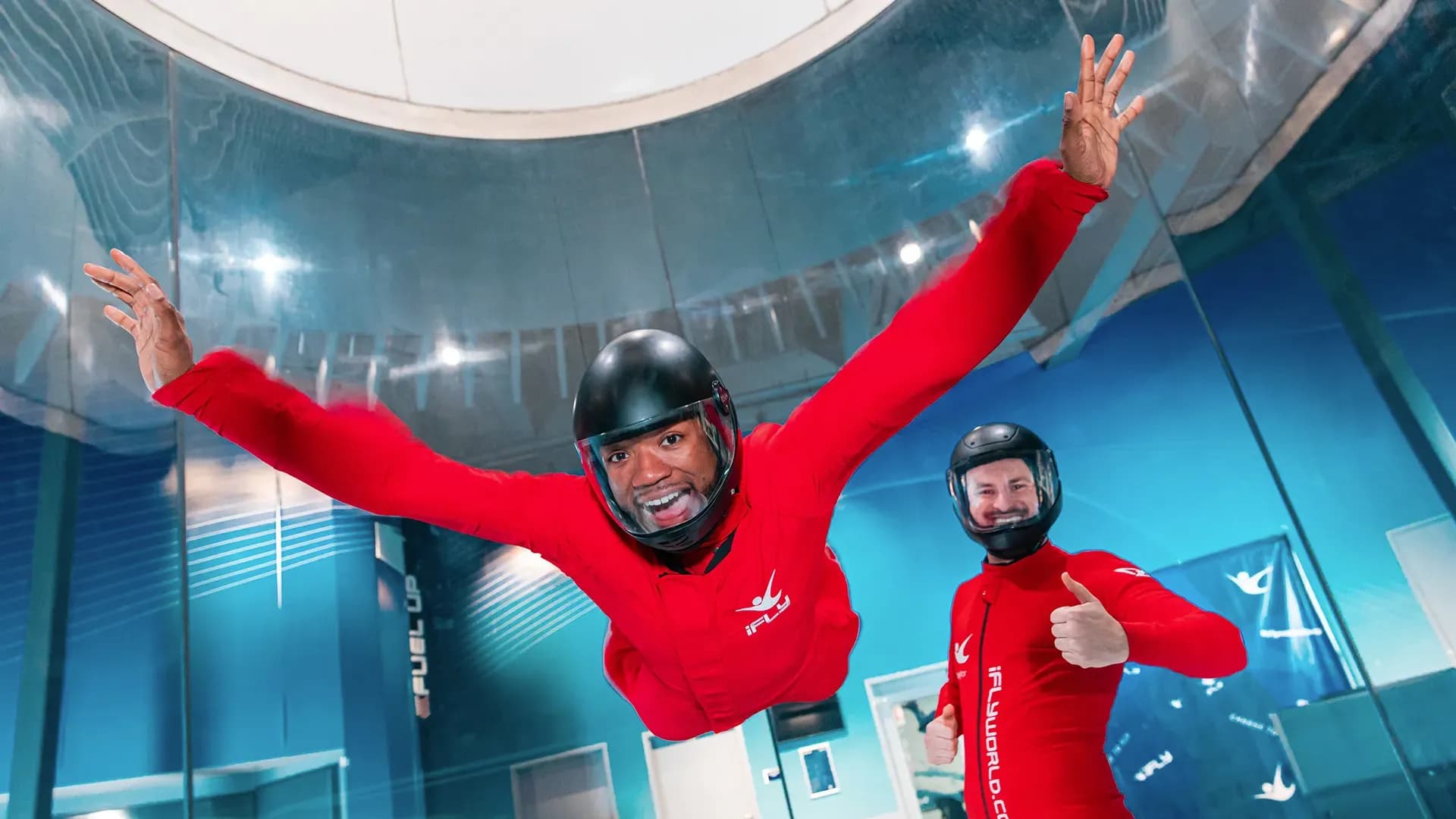 Smiling man in red suit wind tunnel flying, instructor gives thumbs up.