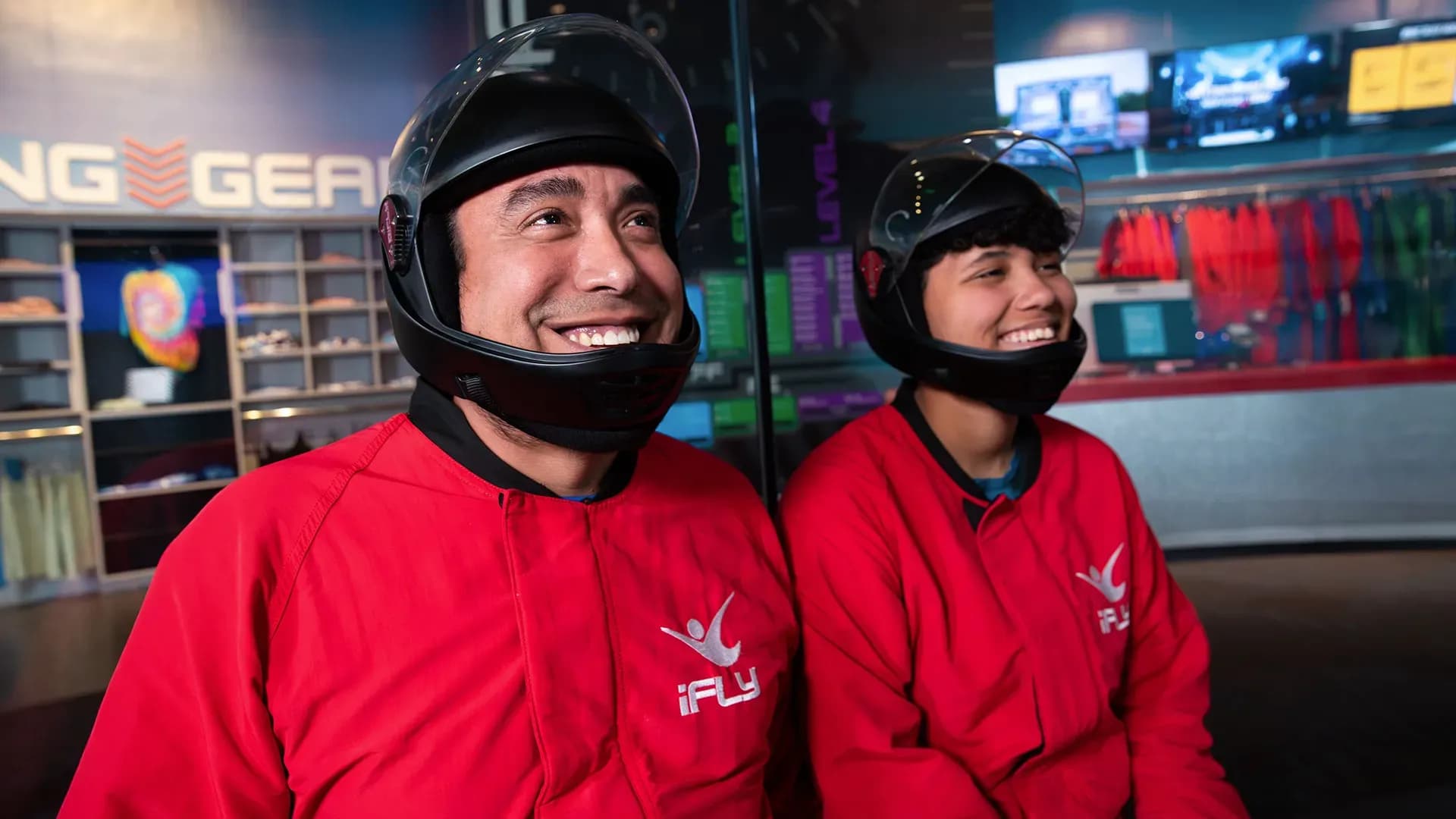Two smiling people in red jumpsuits and black helmets prepare for indoor skydiving.