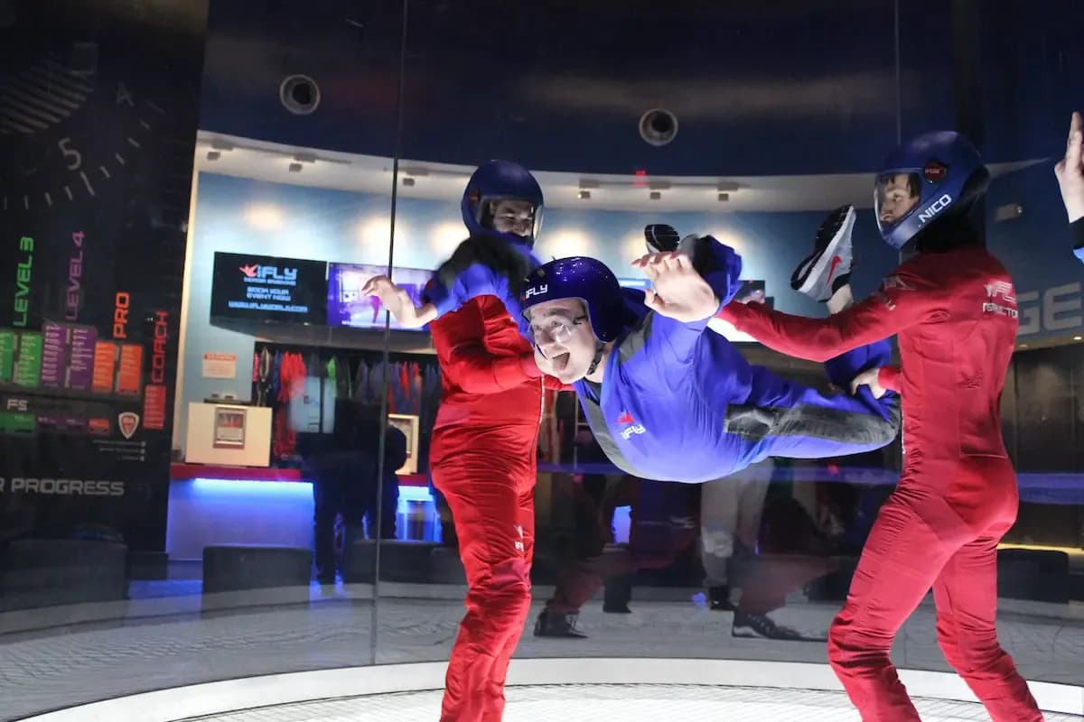 Three people in specialized suits and helmets experience indoor skydiving with instructors.