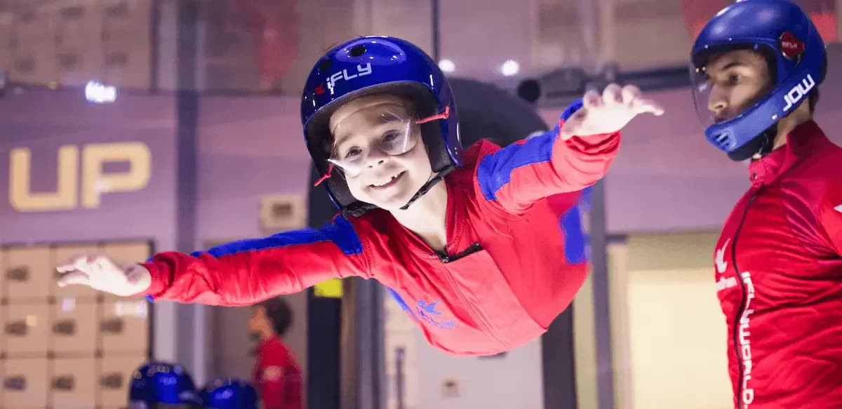 Smiling child in red flight suit floats happily in an indoor skydiving wind tunnel.