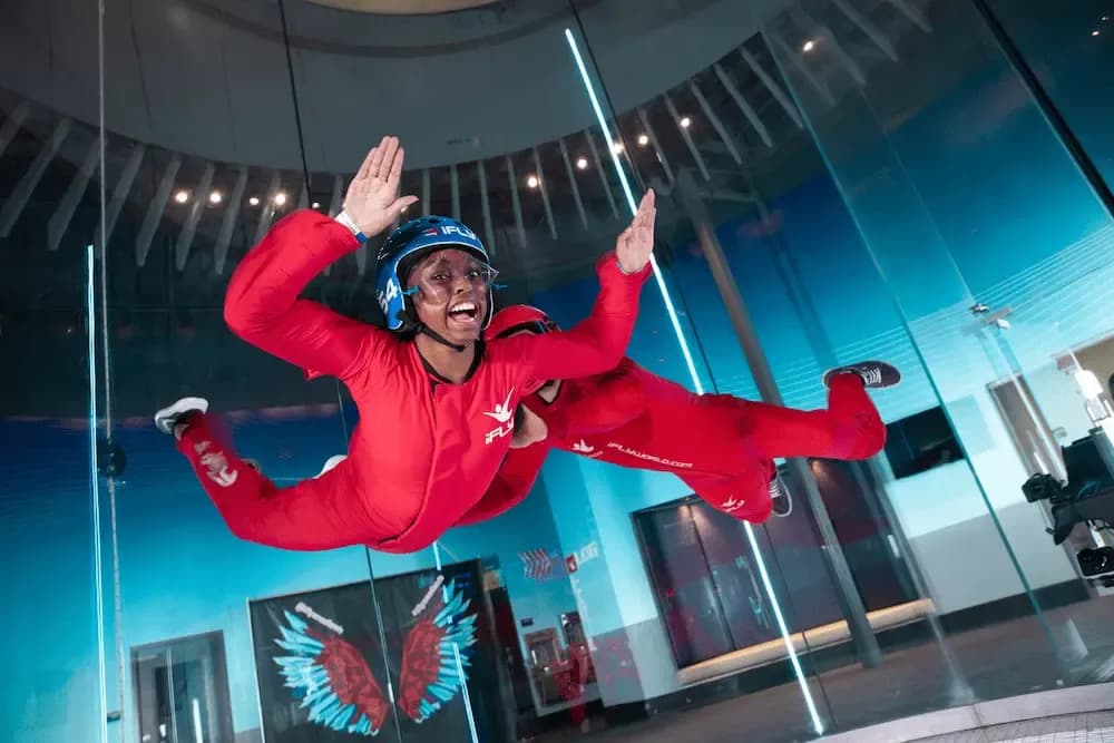 Smiling person in red jumpsuit indoor skydiving, hands raised, with an instructor.