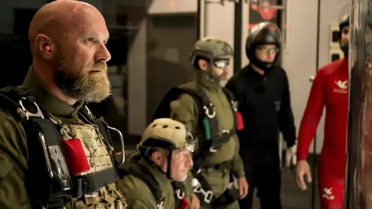 man in military gear with a parachute pack looks right among fellow skydivers.