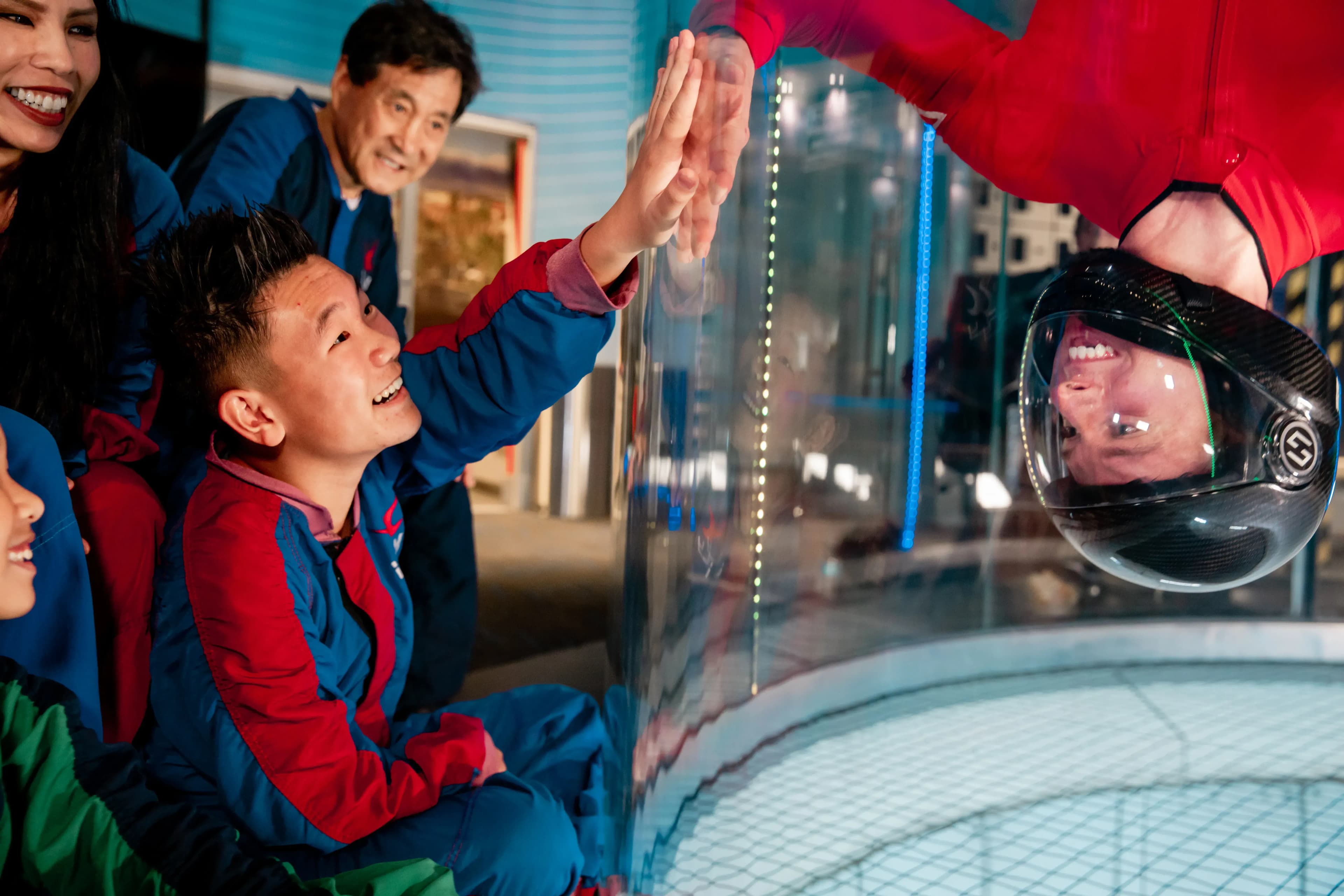 Instructor flying upside down in the tunnel place his handle against the glass, while a young boy outside the tunnel also places his hand against the glass