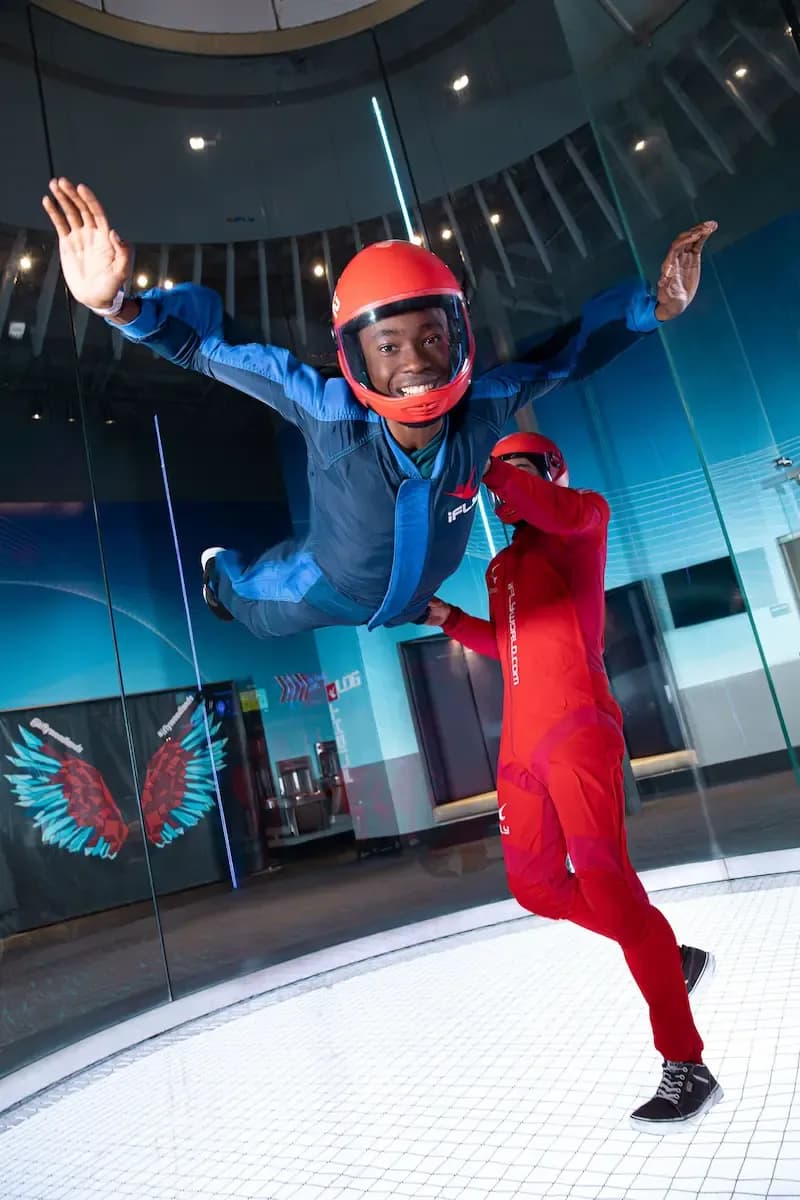 Person in blue suit and helmet indoor skydiving, guided by an instructor in red.