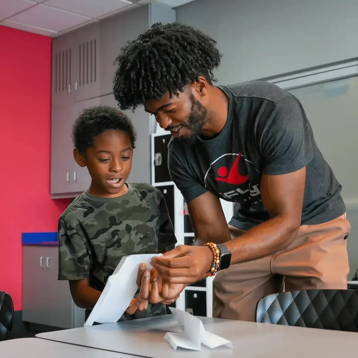 An adult male guides a young boy using a white tablet in a classroom setting.