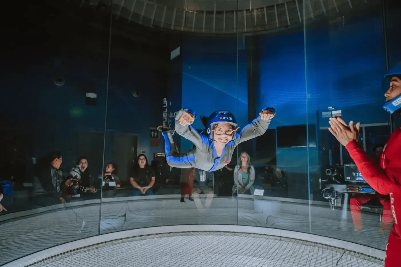 Child in blue flight suit smiles while indoor skydiving, guided by an instructor.