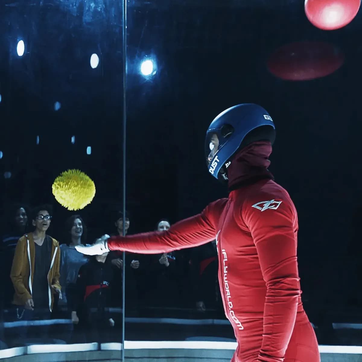 Person in red suit and helmet reaching for a yellow ball floating in a wind tunnel.