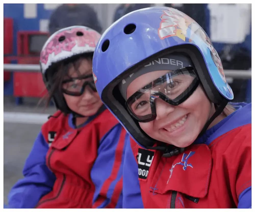 Two young children in helmets and goggles, one smiling, ready for a race event.