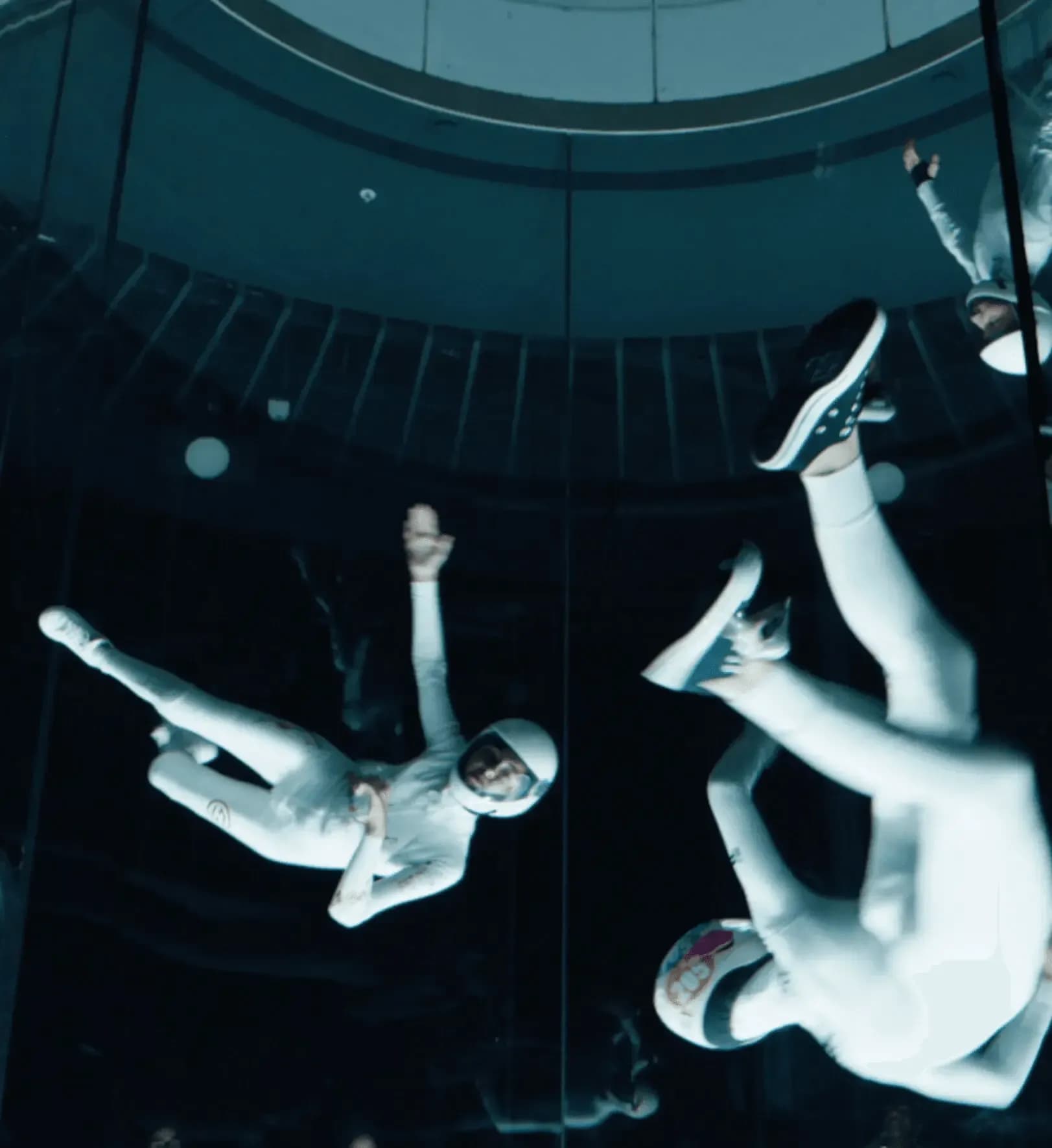 Two people in white jumpsuits indoor skydiving in a dark, vertical wind tunnel.