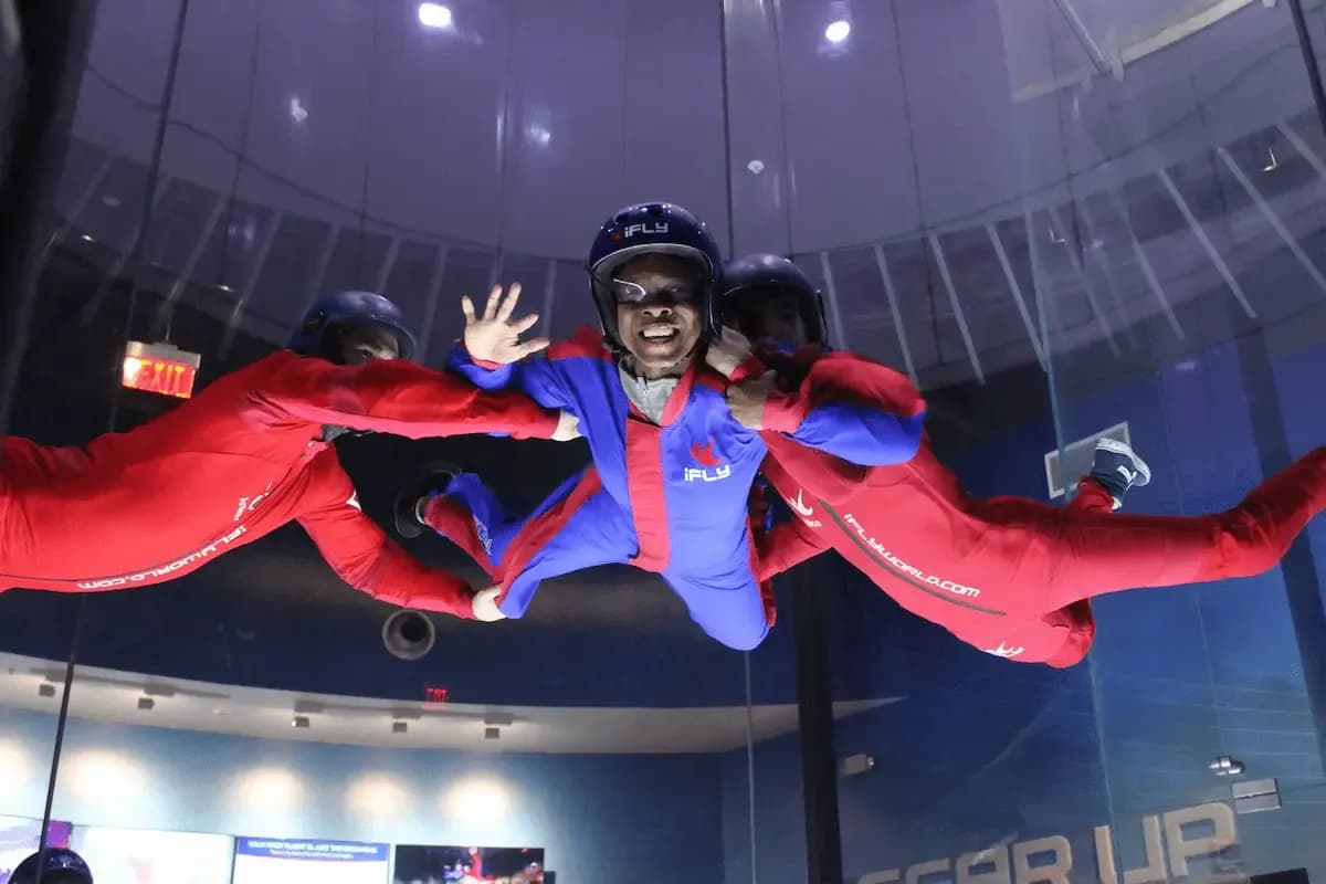 A person in a blue and red jumpsuit smiles while indoor skydiving in a wind tunnel.