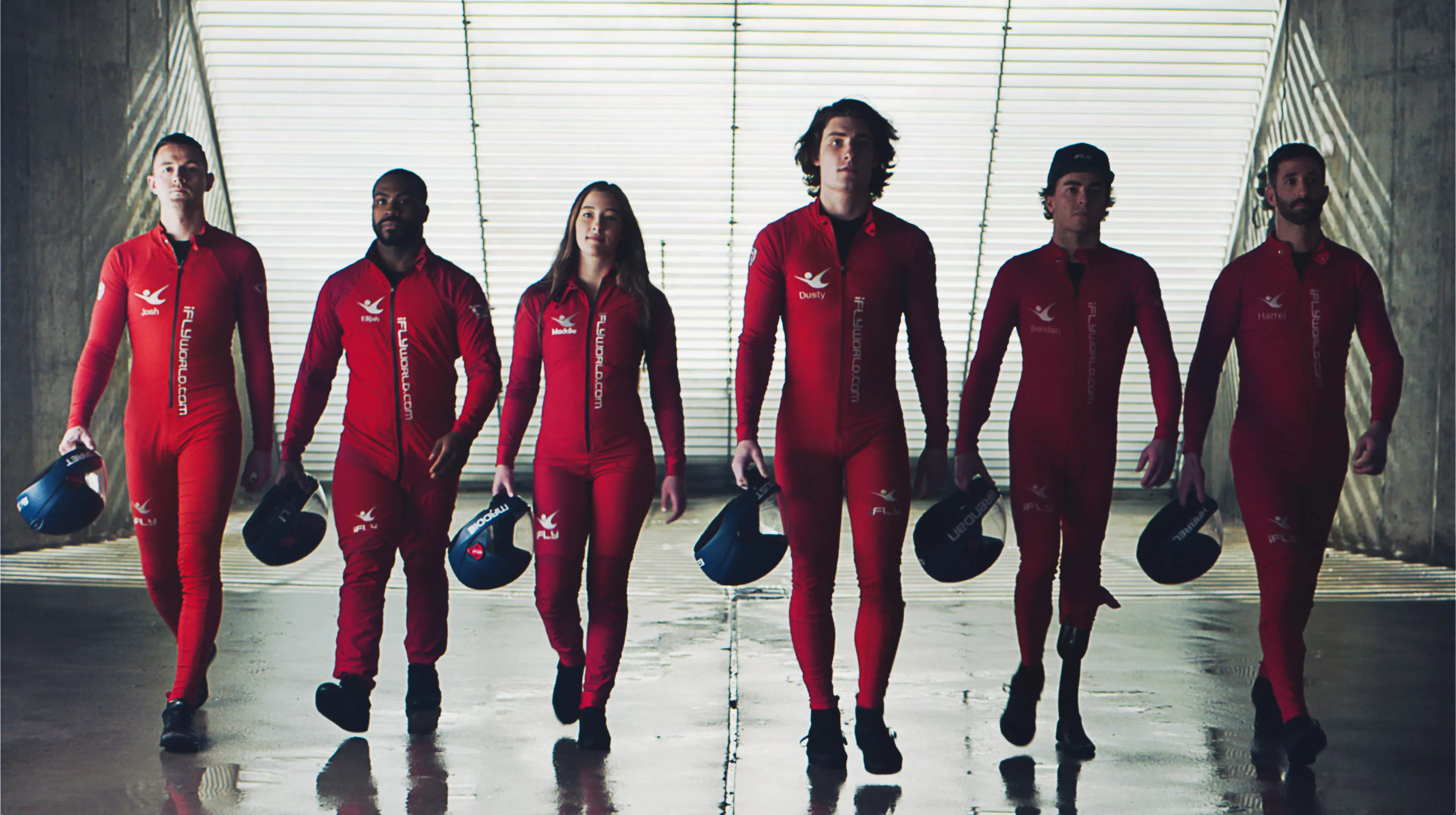 Six young adults walking in a line wearing red iFLY skydiving suits and holding blue helmets