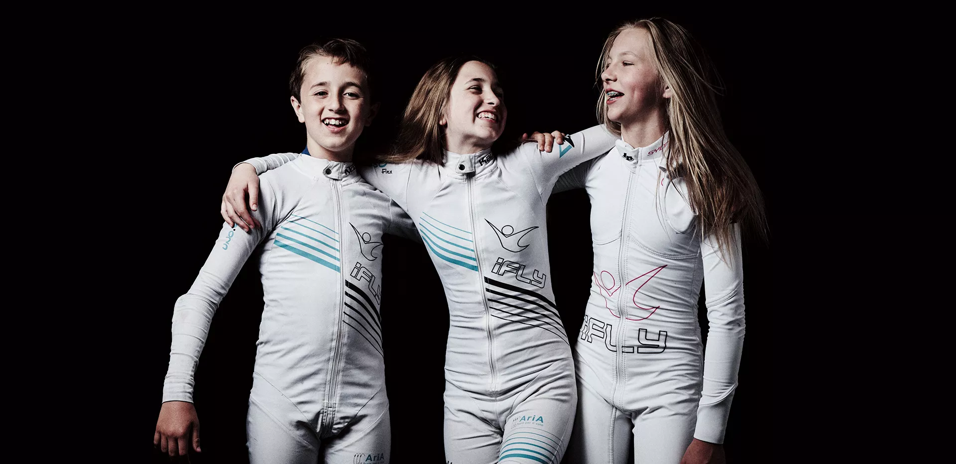 Three children wearing iFLY suits laughing with their arms around one another
