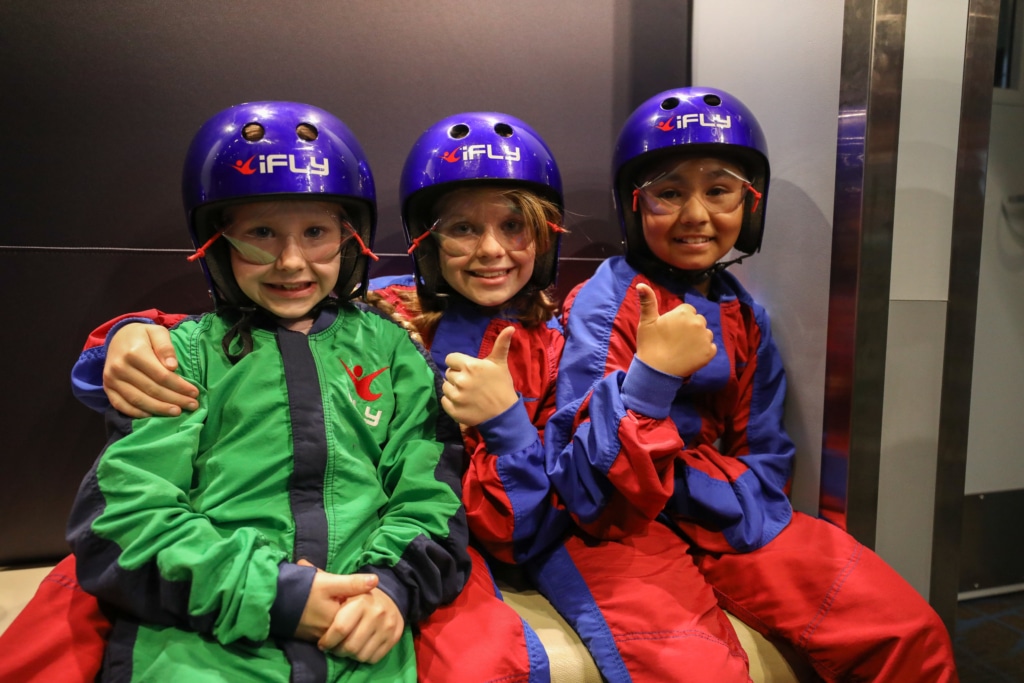 Three children wearing iFLY suits laughing with their arms around one another
