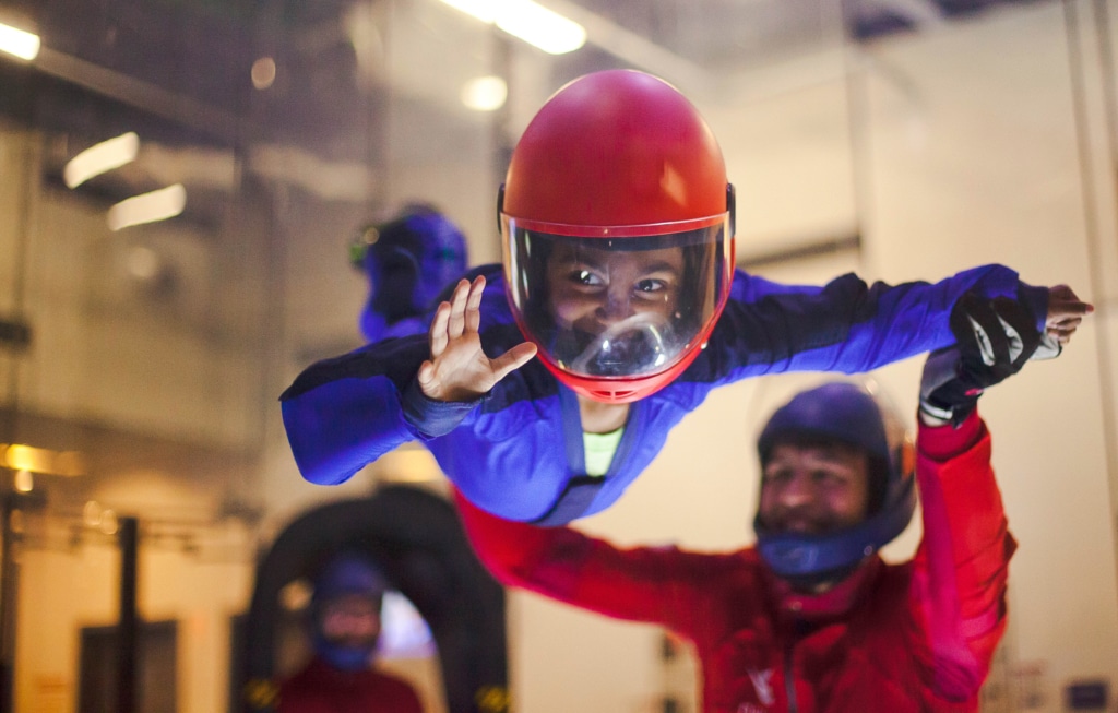 Indoor skydiving instructor helping a child fly in tunnel with an audience around watching