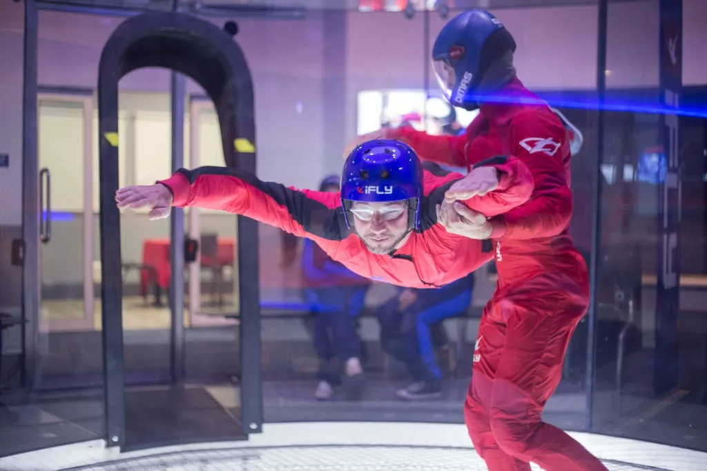 Indoor skydiving instructor helping an adult fly in tunnel with a group of people around watching